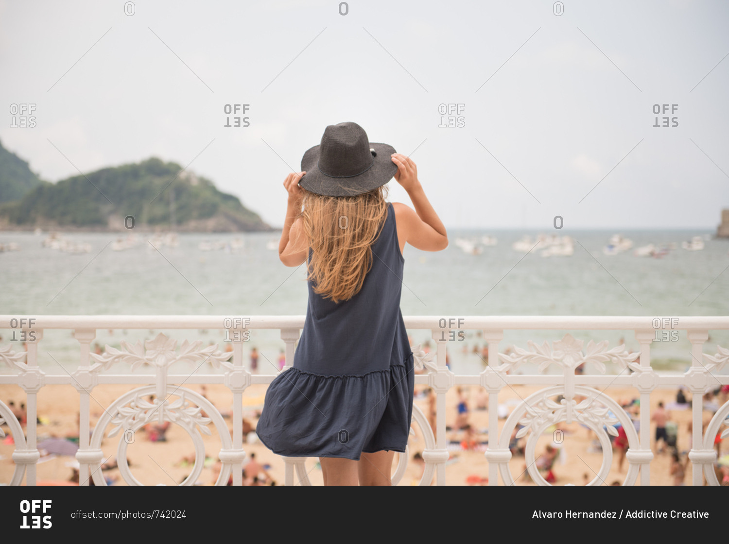 Back view of young lady holding hat and enjoying view of crowded beach and calm sea while standing near fence