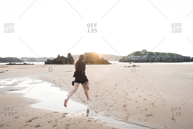 Unrecognizable barefoot lady holding hat and footwear while jumping over stream on sandy beach