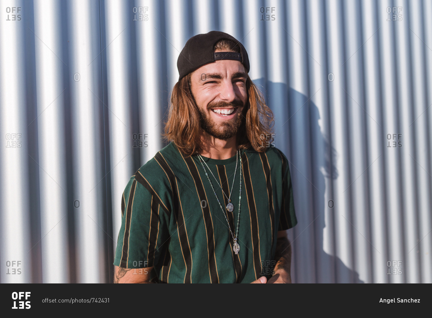 Front view of young man with beard long hair and cap using his mobile phone  in the city on a sunny day of summer stock photo  OFFSET
