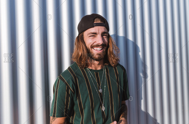 Front view of young man with beard long hair and cap smiling in a sunny day  iron wall background stock photo - OFFSET