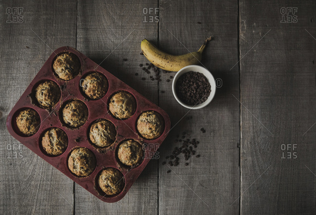 High angle view of baked muffins in baking sheet by chocolate chips and banana on wooden table at home