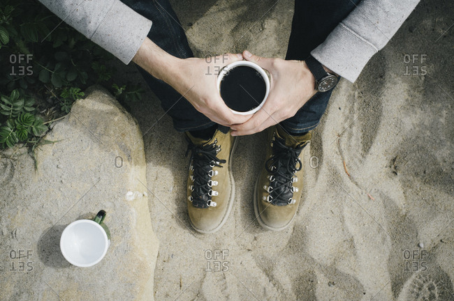 Low section of man holding coffee cup while sitting on sand at beach