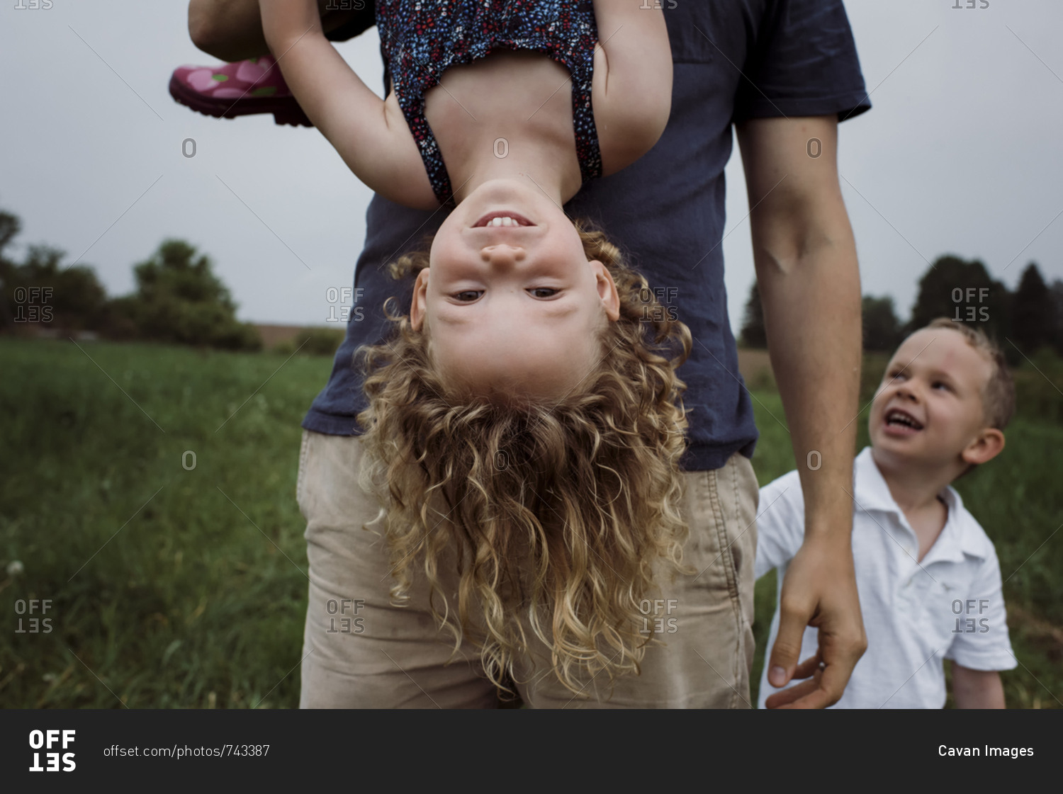 Midsection of father picking daughter upside down while standing with son on grassy field against sky at park