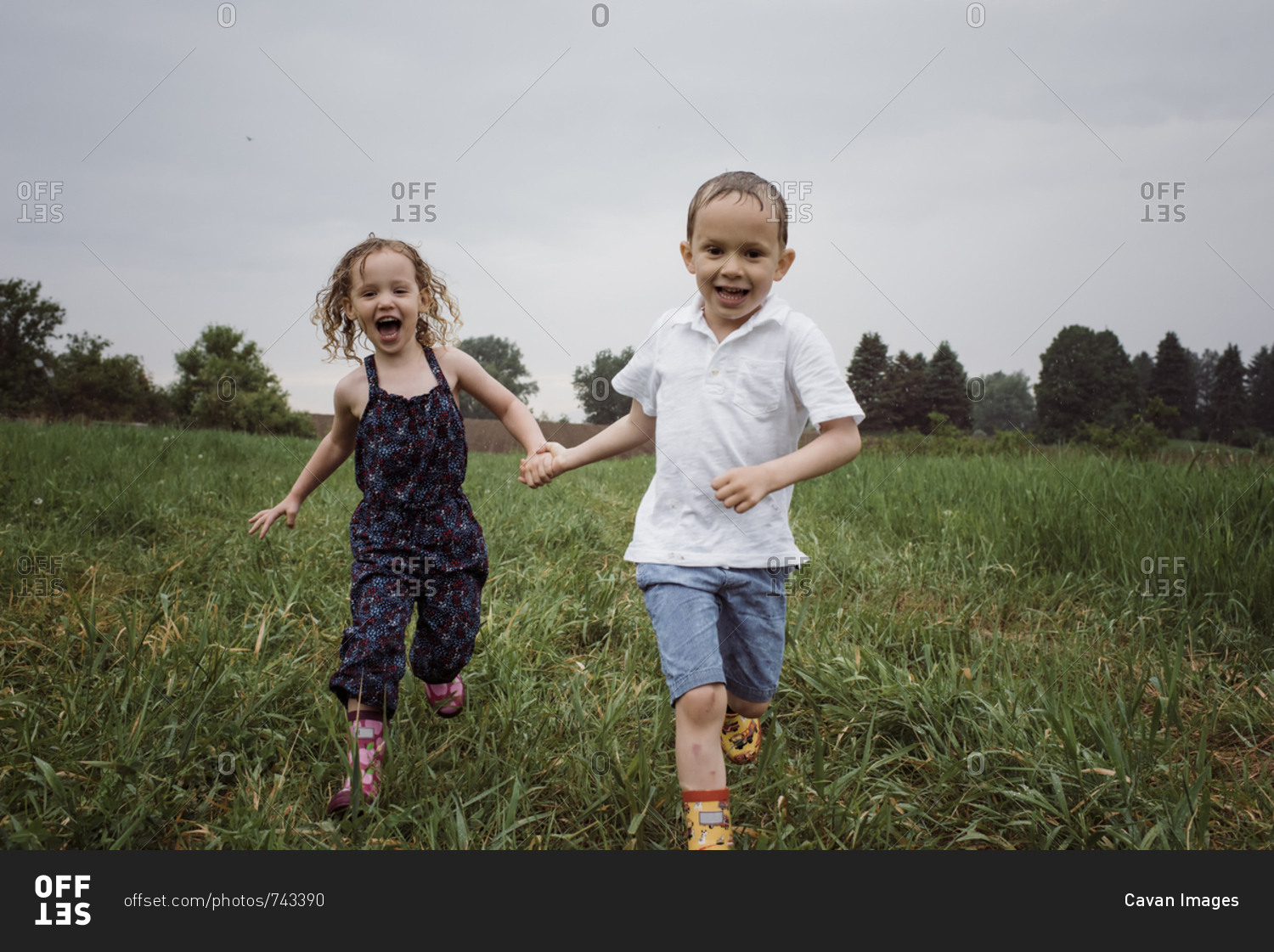 Portrait of happy wet siblings holding hands while running on grassy field against sky at park during rainy season