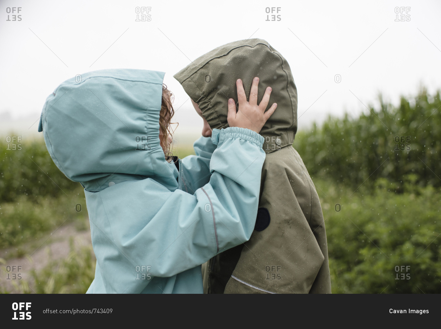 Side view of siblings in raincoats looking at each other while standing against plants during rainy season