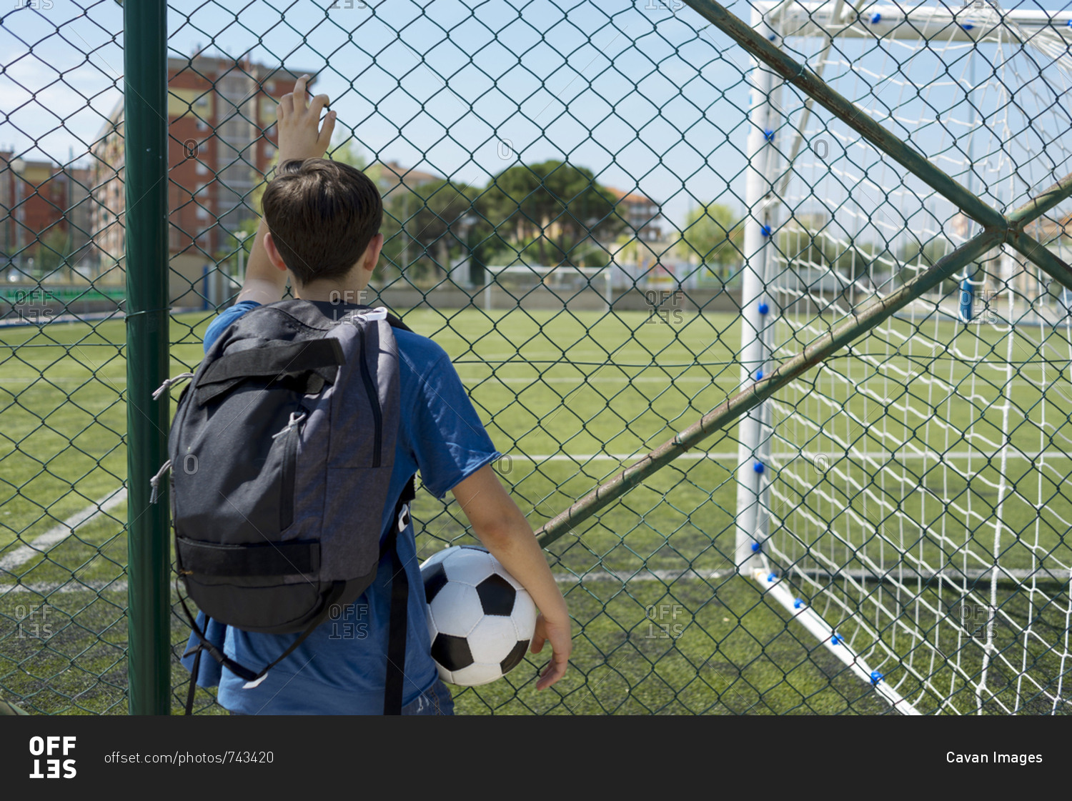 Rear view of boy with backpack holding soccer ball while standing by fence at dugout