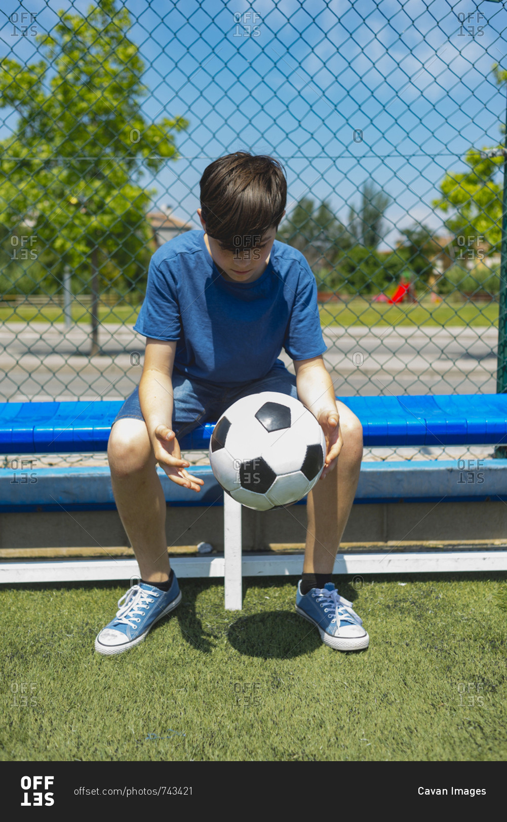 Boy holding soccer ball while sitting on bench by fence at dugout