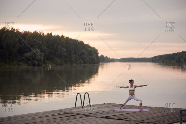 Woman practicing warrior 2 pose on pier by lake against cloudy sky during sunset