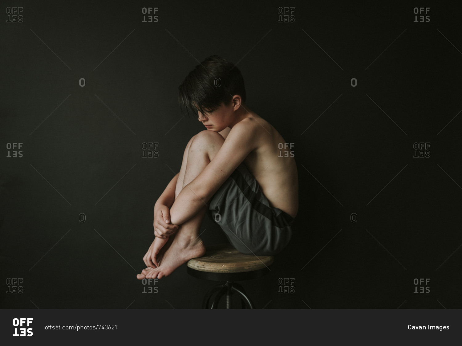 Side view of sad shirtless boy sitting on stool against black background