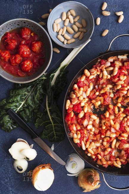 Tomatoes, onions and cannellini beans in a pan beside silverbeet, tomatoes and sliced onions.