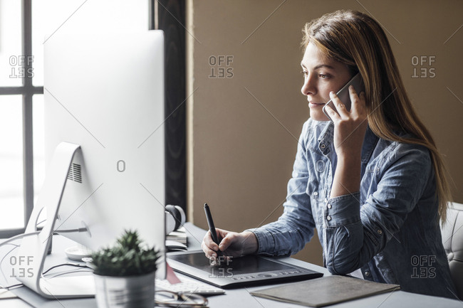 Beautiful blonde Caucasian businesswoman graphic designer working on her computer and talking on cell phone.
