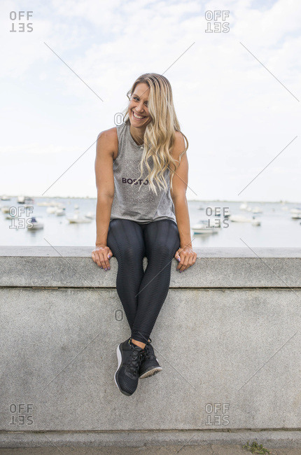 Smiling Young Adult Woman in Fitness Attire Sitting on Stone Wall with Ocean in Background