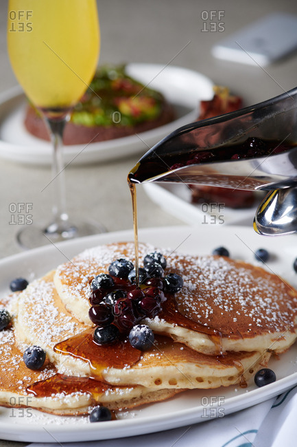 Blueberry Pancakes with syrup being poured over the blueberries and dripping onto the pancakes below. In the background is avocado toast, a stack of chopped bacon and a mimosa.