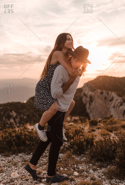 Young blond vintage man with black hat carrying his girlfriend piggyback in a cliff sunset and mountains background