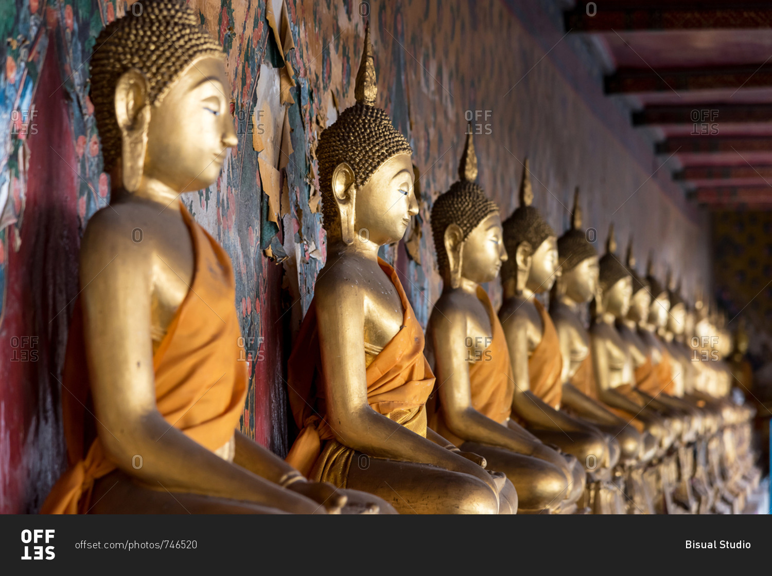 Golden similar statues of Buddha placed in line near patterned wall in Buddhist temple