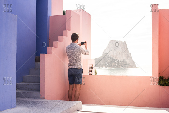 Man seating in a blue and pink building taking a photography of the landscape with a mobile phone