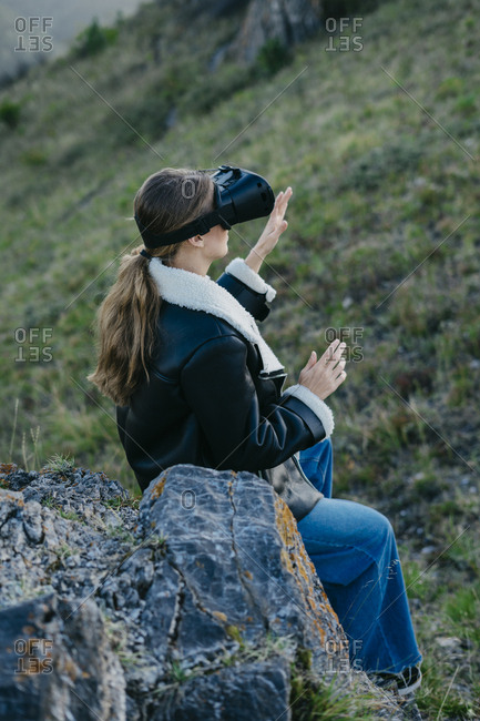 Portrait of a beautiful young woman wearing virtual reality glasses is looking up sitting on a rock