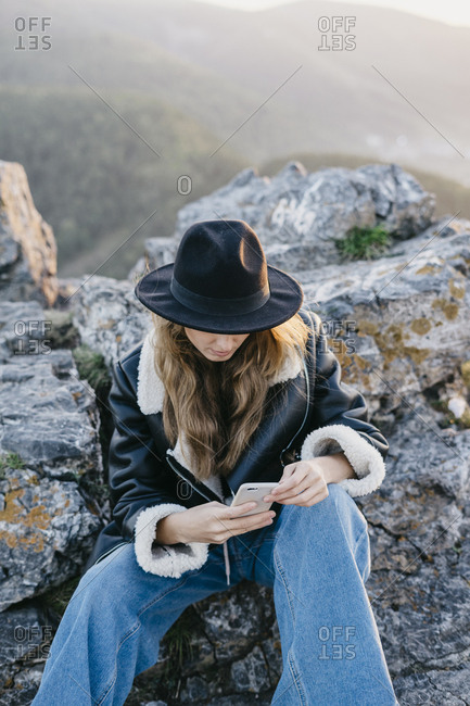 Portrait of a beautiful young woman in a black jacket and hat sitting on a rock