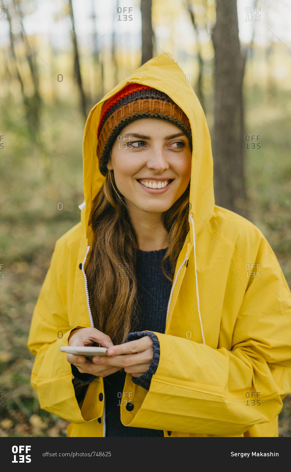 Portrait of a beautiful young woman in a yellow raincoat and knitted hat typing a message on her smart phone
