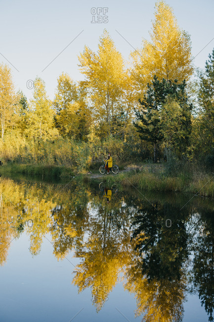 Portrait of a beautiful young woman in a yellow raincoat and knitted hat standing on her bike while looking out at the lake