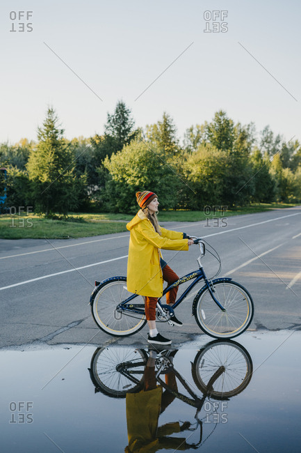 Portrait of a beautiful young woman in a yellow raincoat and knitted hat standing near a large puddle while on her retro bike