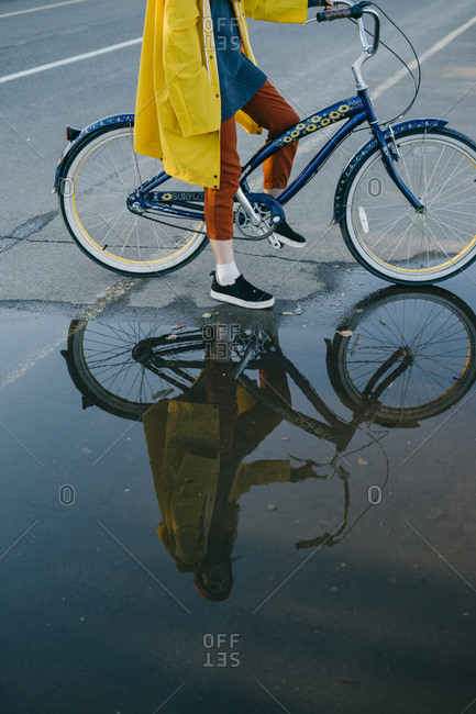 Portrait of a beautiful young woman in a yellow raincoat and knitted hat standing near a large puddle while on her retro bike