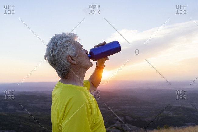 Spain, Catalonia, Montcau, senior man drinking water and looking at view from top of hill during sunset