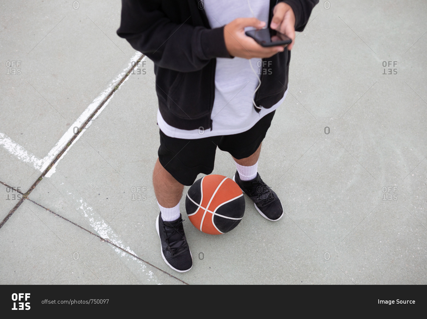 Male teenage basketball player standing on basketball court looking at smartphone, waist down