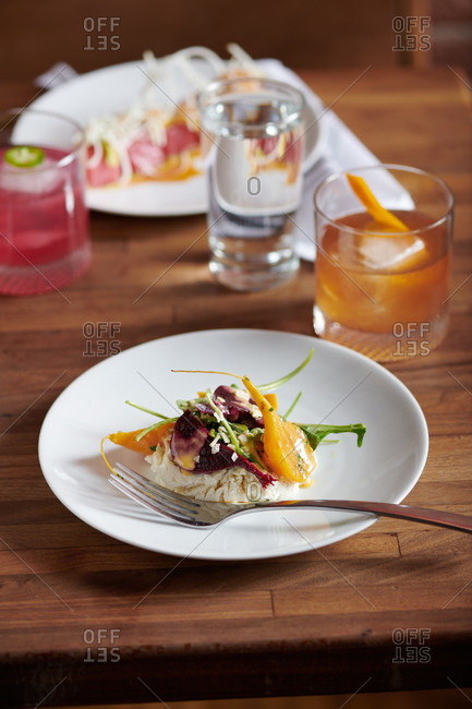 An elegant beet salad on a wooden table.  The salad consists of beets, arugula, mandarins, goat cheese, candied fennel, and honey.  Nearby a Fig Old Fashioned cocktail of rye whiskey, fig puree angostiura and old fashioned bitters with an orange peel twist.