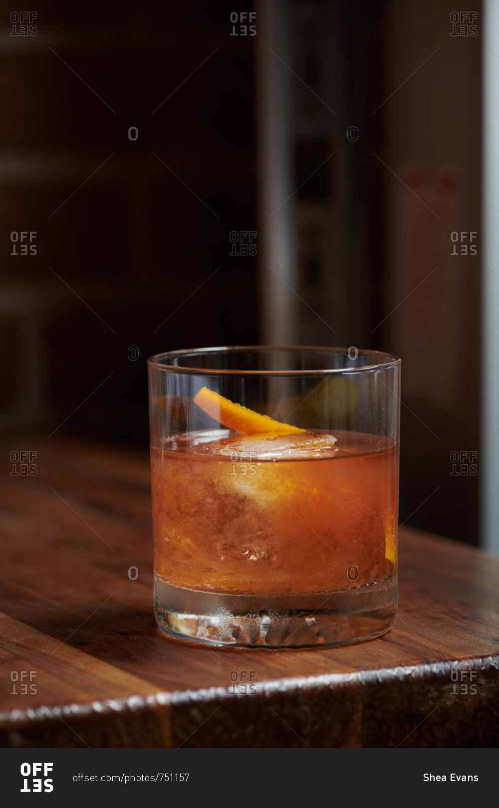 An old fashioned cocktail sits on a wooden table in a tapas style restaurant with a twist of orange peel garnishing the rocks glass with a single ice cube inside. The drink consists of rye whiskey, fig puree, angosturia, and old fashioned bitters.