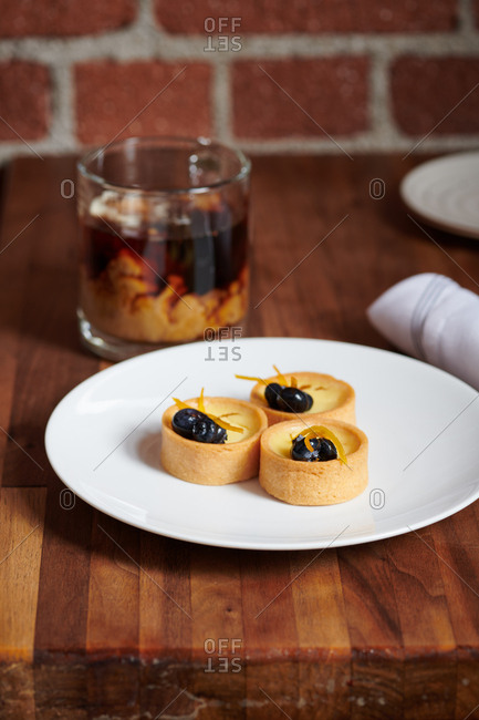 Three Lemon Tartlets served on a white plate with blueberries and candied orange peels for dessert in a Tapas style restaurant.  Nearby a coffee with just added cream sits to accompany the dessert.