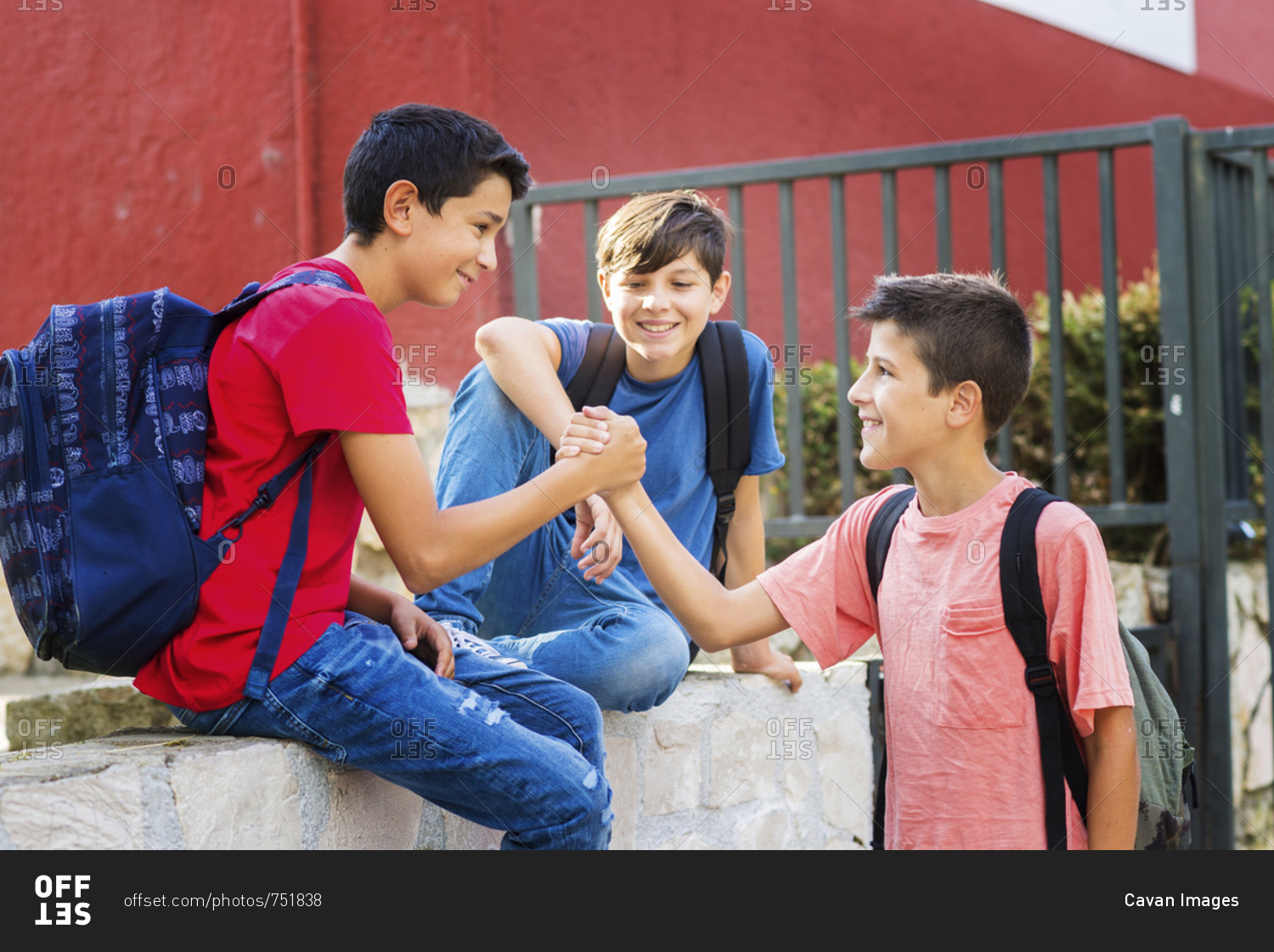Smiling boy looking at friends giving handshake while sitting on retaining wall against school building