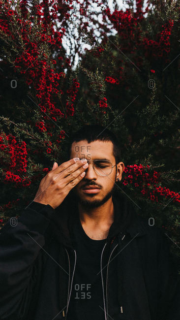 Handsome man in glasses covering eye with hand while standing near tree with pretty red flowers