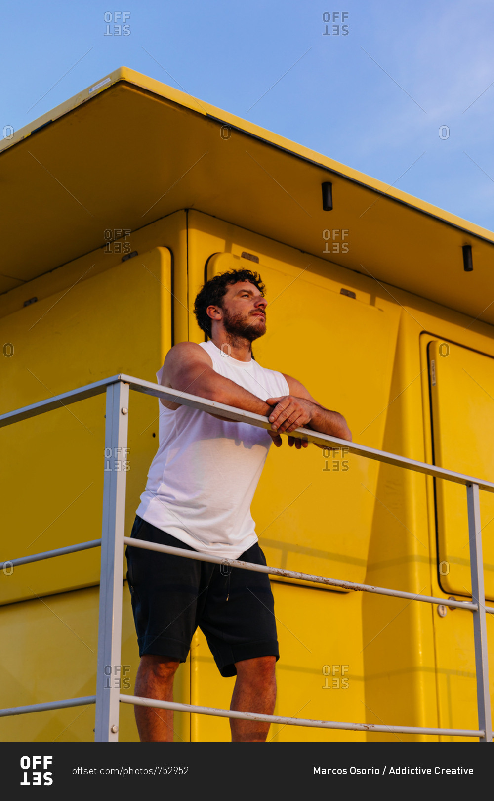 Handsome bearded man in sportswear leaning on railing of lifeguard cabin on beach during outdoor workout