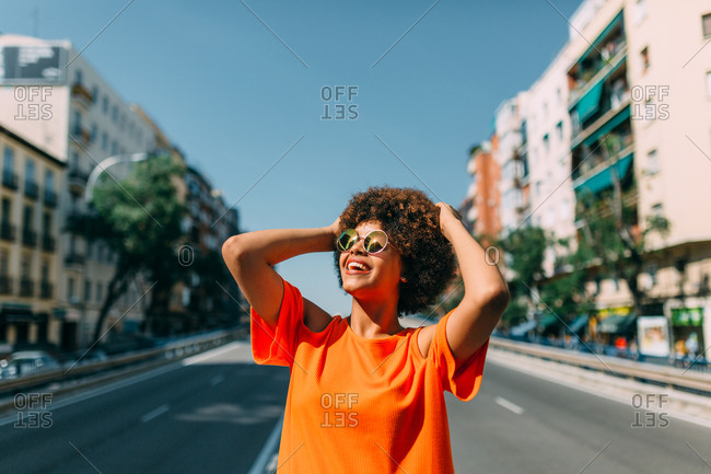 Lovely African-American woman in sunglasses cheerfully smiling and looking at clear blue sky while standing in middle of road in nice city