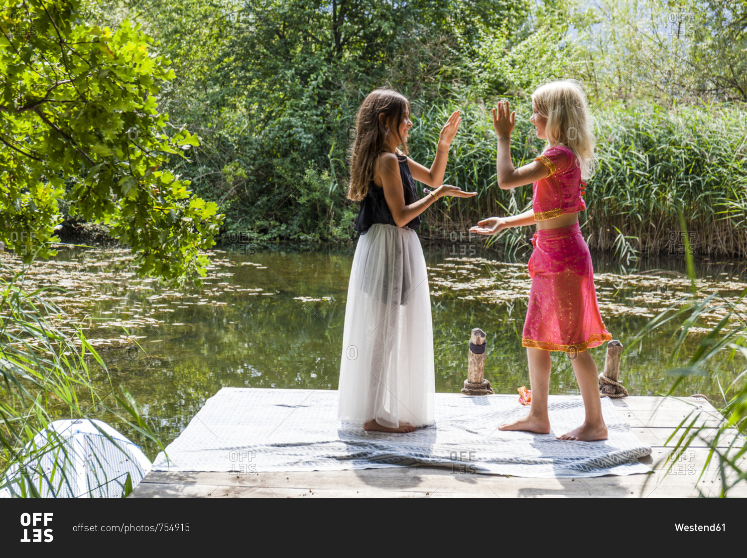 Two girls standing on jetty at a pond in fancy dresses doing a hand clapping game