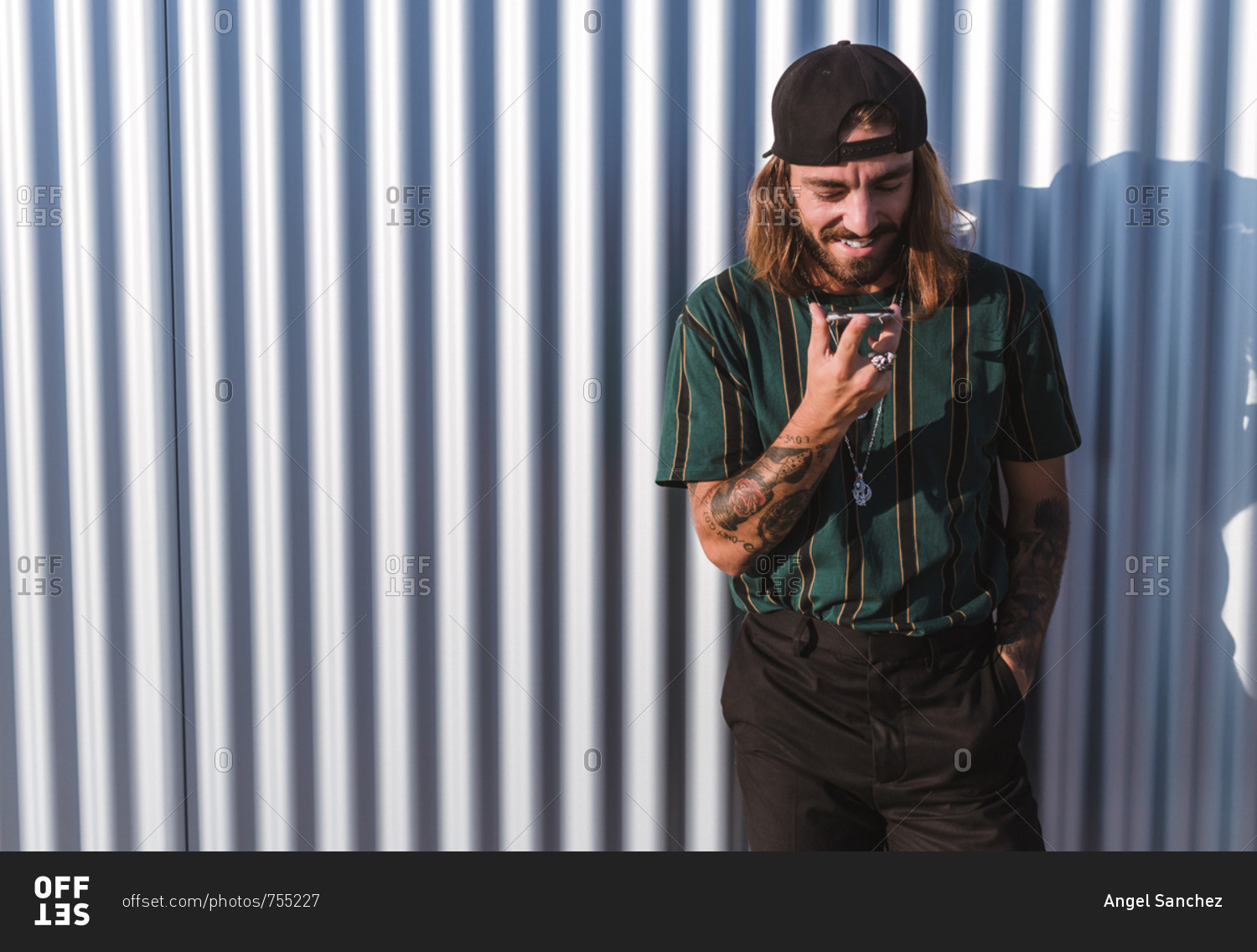 Front view of young man with beard long hair and cap using his mobile phone  in the city on a sunny day of summer stock photo - OFFSET