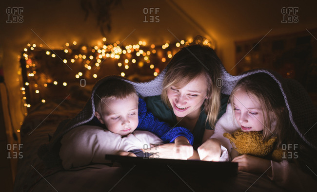 Mother and kids under the blanket using digital tablet against Christmas lights