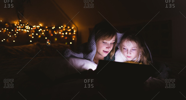 Mother and daughter under the blanket using digital tablet against Christmas lights