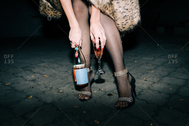 Woman in faux fur coat and high heels holding a bottle and a glass of champagne outside at night