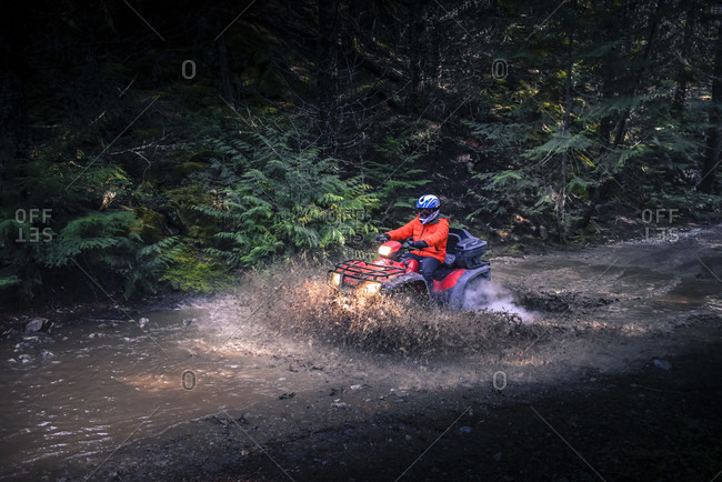 February 15, 2015: Side view of adventurous man driving quadbike through mud on dirt road in forest, Callaghan Valley, Whistler, British Columbia, Canada