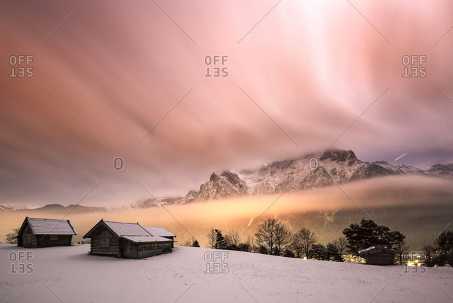 Night photography of the Karwendelgebirge (mountains) in winter, with snow and wooden hut in the foreground. Nebulous mood by the lights of the market Mittenwald illuminated, while the clouds seem strip-type over the mountains by the long time exposure.