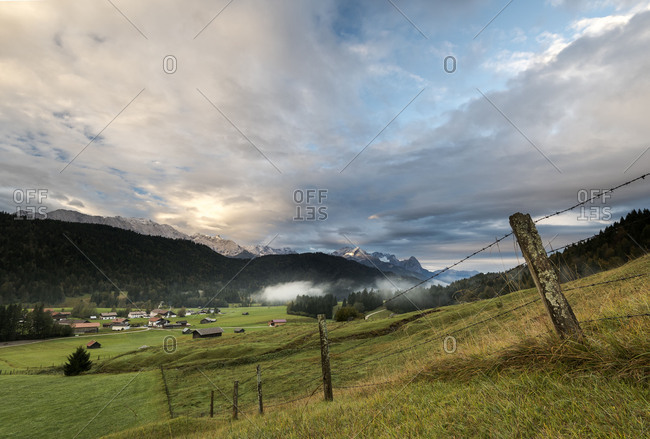 mossy pasture fence in a mountain pasture, in the background the hamlet Gerold, fog and clouds, as well as the Zugspitze massif in the morning light.