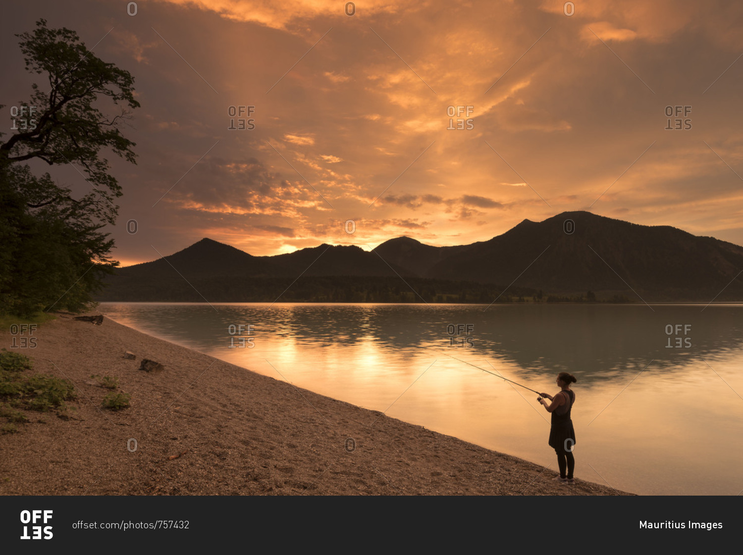 Young woman fishes on the shore of the Walchensee / Lake Walchen while dramatic sundown