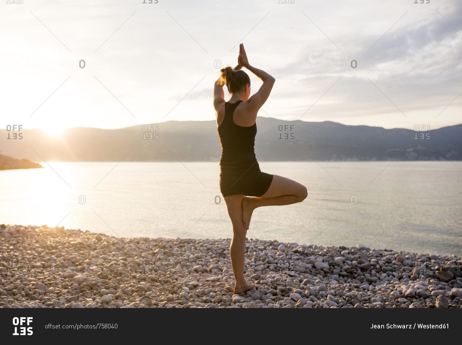 Young woman doing yoga on a stony beach at sunset- tree position