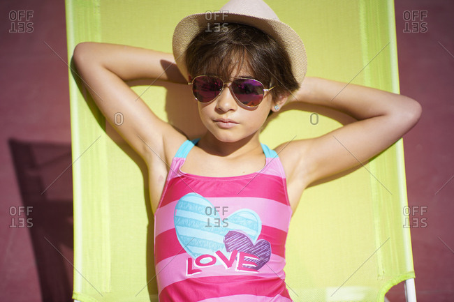 Young girl wearing swimwear- sunglasses and a sun hat- sitting in sun lounger