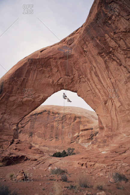 Rock climber hanging under eroded natural arch, Moab, Utah, USA