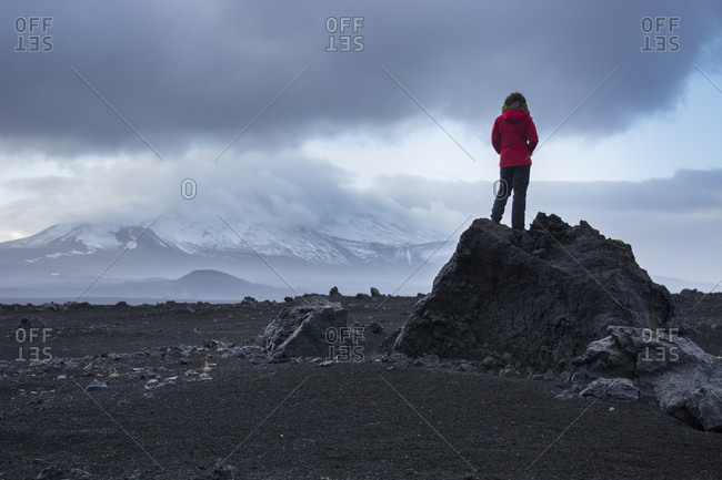 A young adventurous woman with a red jacket, stands on top of a black, volcanic rock as she stares off into the distance, gazing at the snowy mountain covered by clouds, Hekla, in Iceland