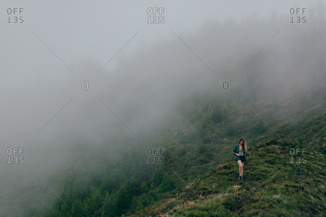 Woman walking on high steep hill covered with green grass with thick fog above