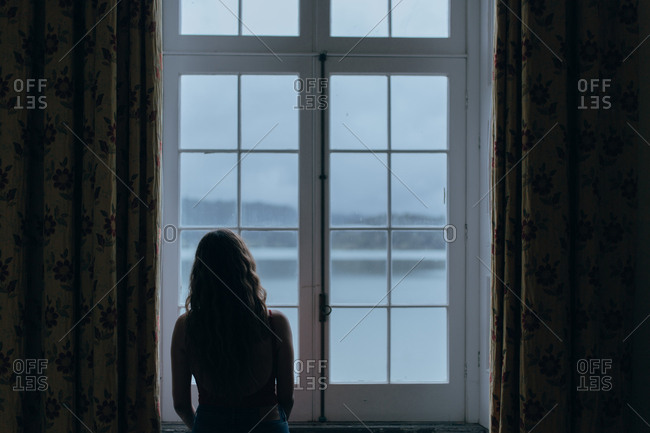 Back view of woman standing near window and looking at quiet lake and mountains under clear sky